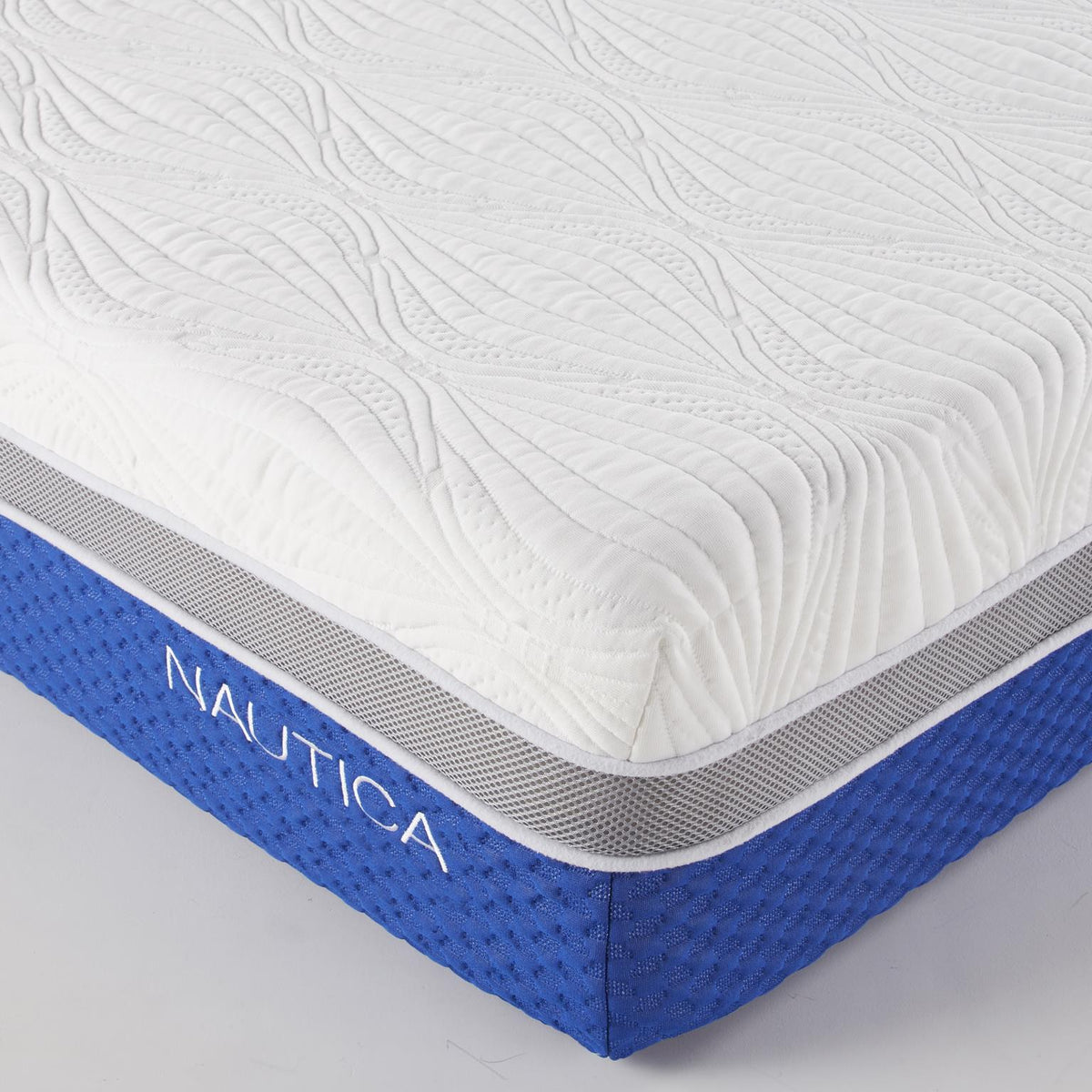 A close-up studio shot of a mattress corner showing a white quilted top surface, a gray mesh band, and a blue side panel with the brand name NAUTICA in white.