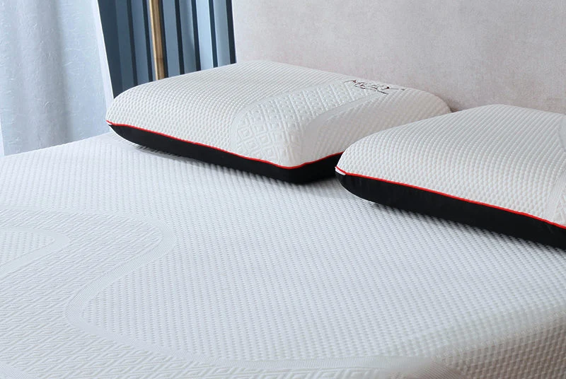 Two white textured pillows with black edges and a thin red piping accent, resting on a white quilted mattress in a bedroom setting.