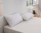 Two white pillows with light blue piping trim resting on a white quilted mattress in a bright, modern bedroom setting.