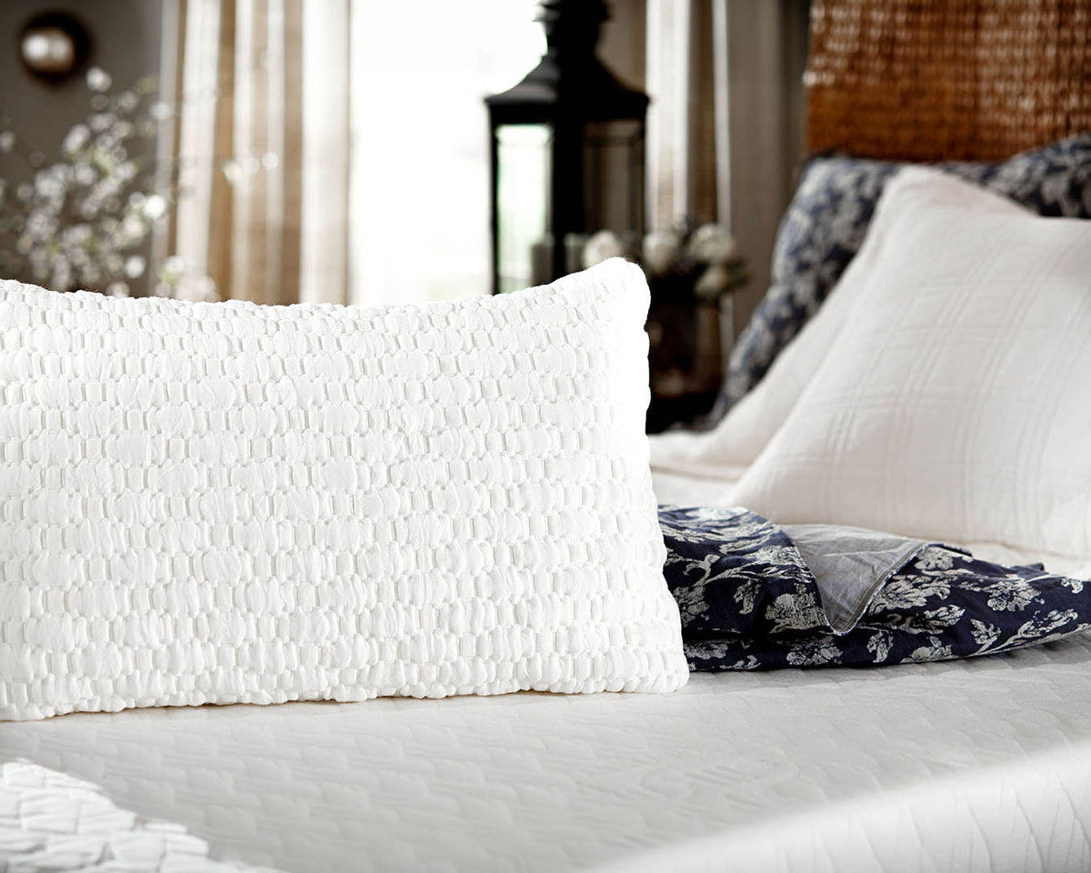 A close-up of a white textured rectangular pillow on a bed, with additional white and navy patterned pillows in a softly lit bedroom setting.