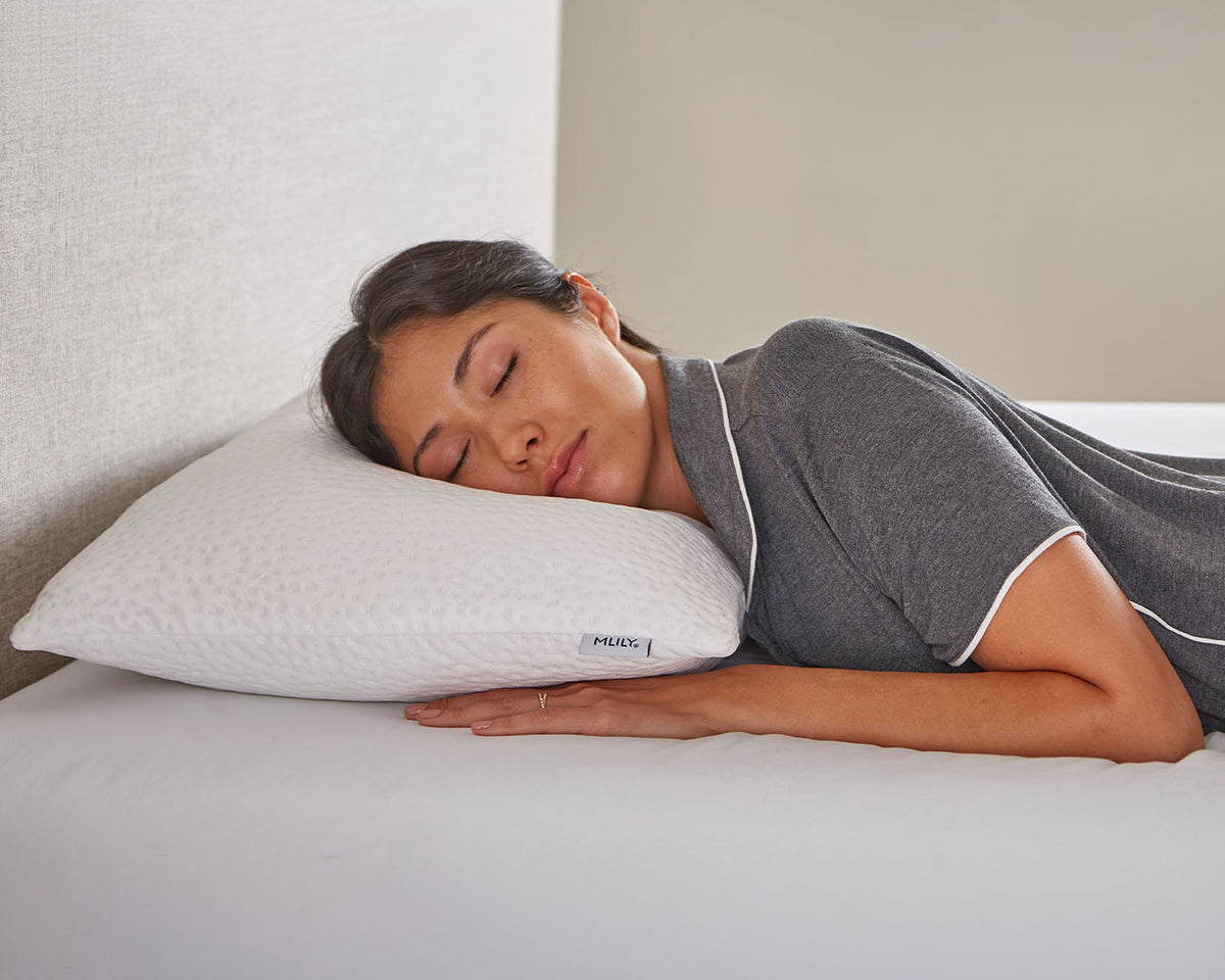 A woman sleeping peacefully on a white textured pillow on a bed, wearing a gray pajama shirt with white piping. The pillow features a small visible brand tag reading 'MORNE'.