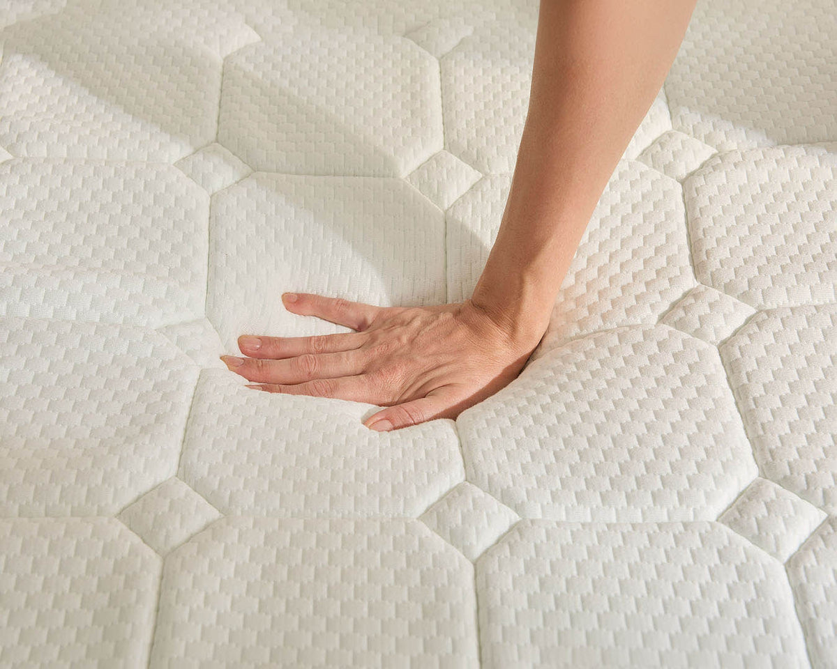 A close-up of a white quilted mattress topper being pressed down by a person's hand and forearm, showing the textured surface and soft indentation.