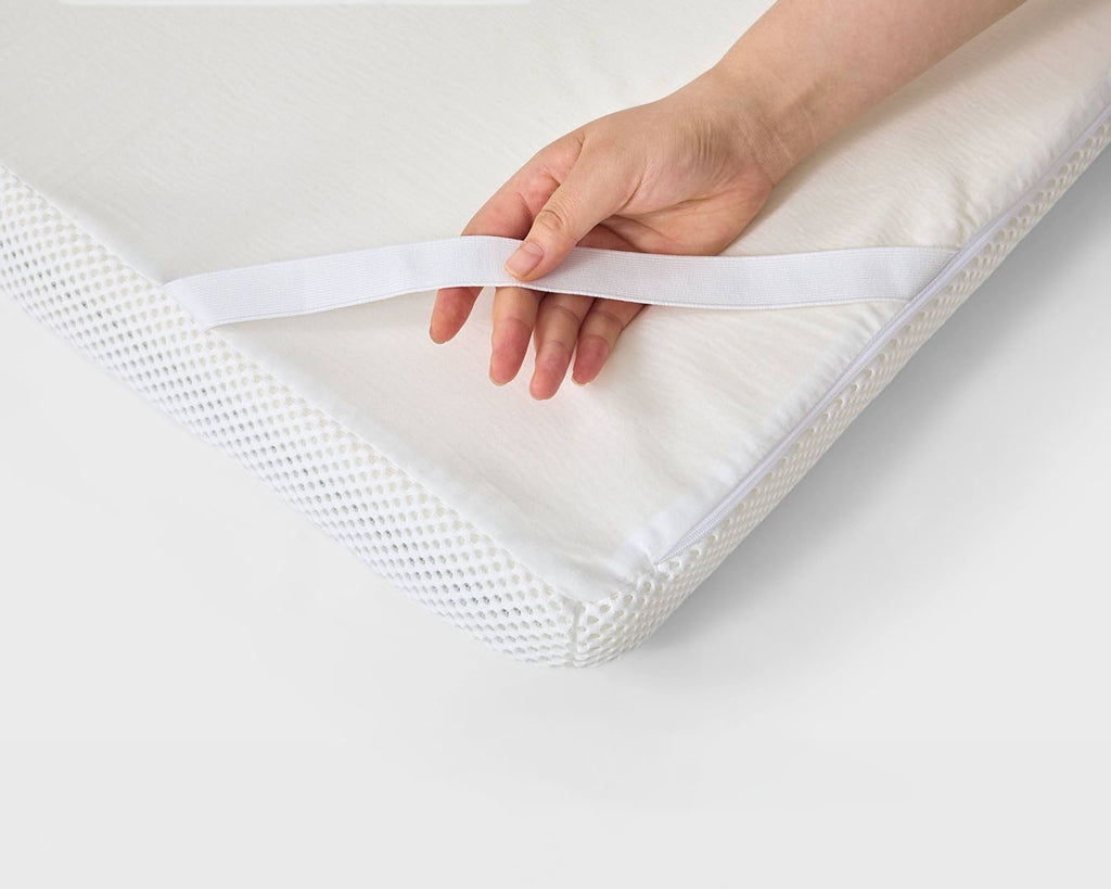 A close-up of a hand pulling a white elastic band off the corner of a white mattress topper or quilted mattress, showing the breathable mesh side of the mattress.