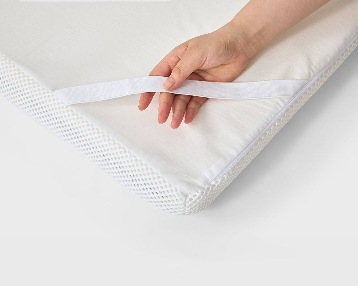 A close-up of a hand pulling a white elastic band off the corner of a white mattress topper or quilted mattress, showing the breathable mesh side of the mattress.