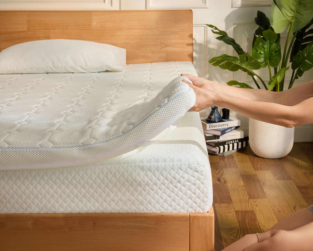 A person lifts a corner of a white quilted mattress topper on a wooden bed frame in a bright bedroom, with a plant and books visible in the background.