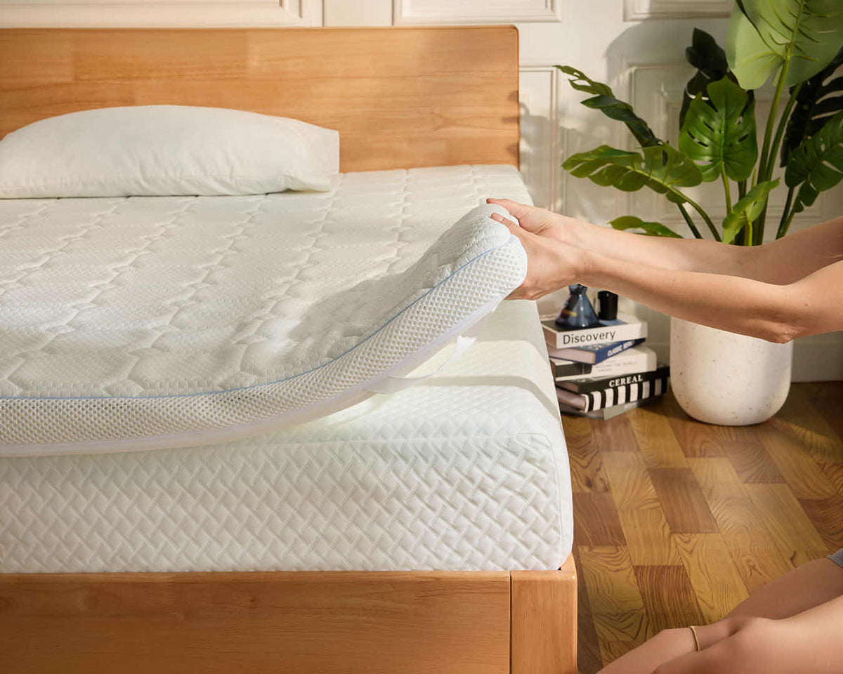 A person lifts a corner of a white quilted mattress topper on a wooden bed frame in a bright bedroom, with a plant and books visible in the background.