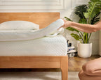 A person lifts a mattress topper off a wooden bed frame in a bright bedroom setting with a potted plant in the background.