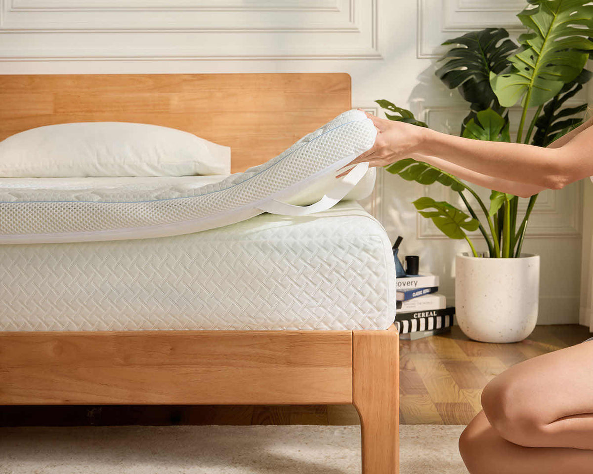 A person lifts a mattress topper off a wooden bed frame in a bright bedroom setting with a potted plant in the background.