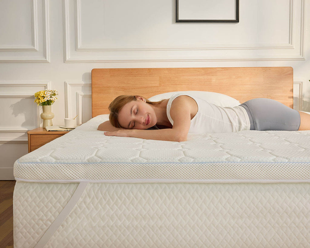 A woman in a white tank top and gray shorts rests on her side on a white quilted mattress topper on a bed with a light wood headboard in a bright bedroom.