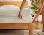 A person's hands are shown peeling back a white protective film from the side of a white quilted mattress on a light wood bed frame in a bright bedroom setting.