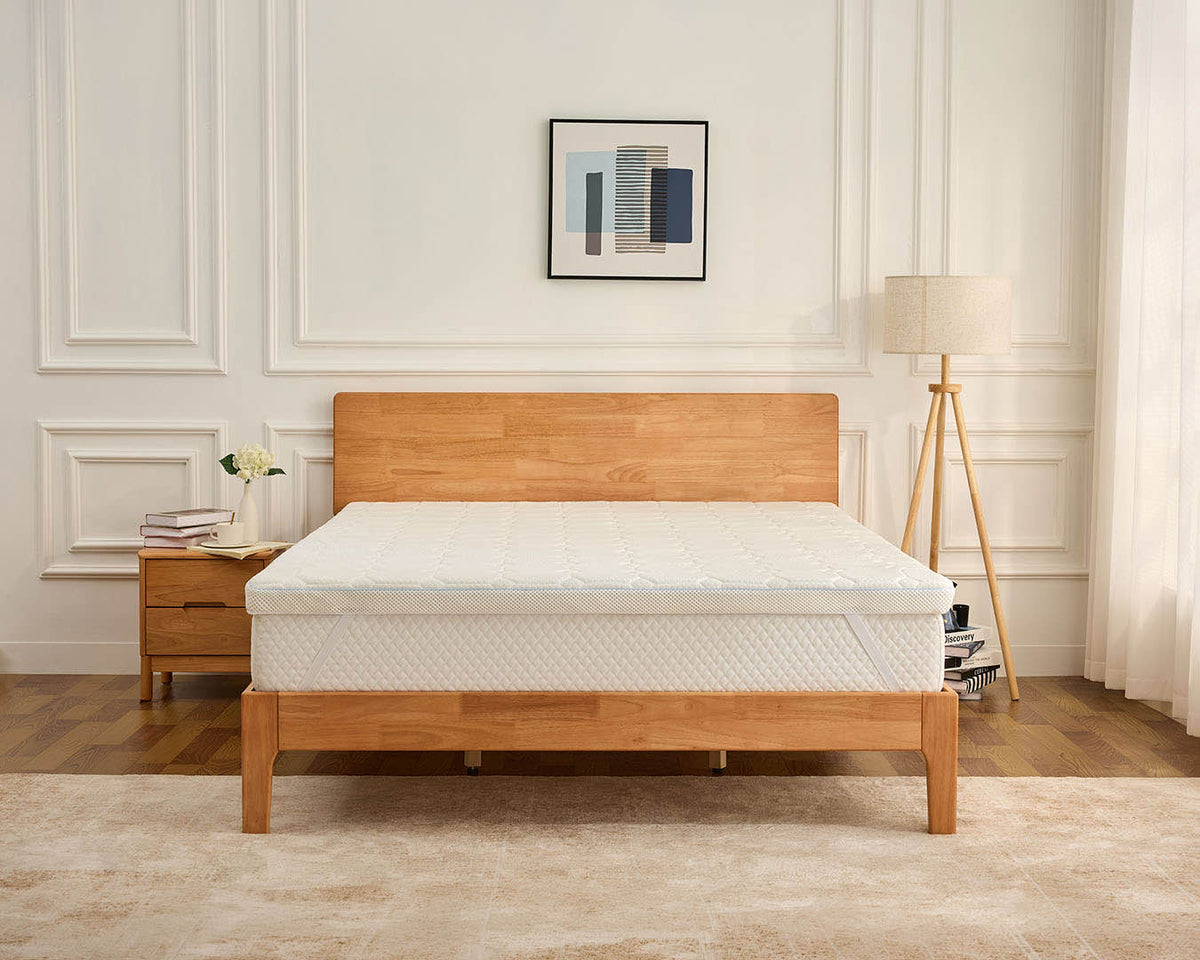 A light oak wood bed frame with a white quilted mattress, styled in a bright bedroom with a matching wooden nightstand, a floor lamp, and a beige rug.