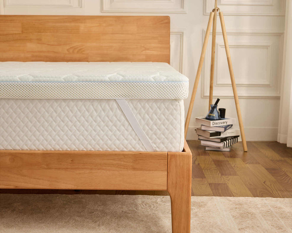 A close-up of a white quilted mattress topper or mattress on a light natural wood bed frame, set in a bright bedroom with a wooden floor, white paneled wall, and a tripod floor lamp.