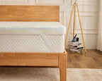 A close-up of a white quilted mattress topper or mattress on a light natural wood bed frame, set in a bright bedroom with a wooden floor, white paneled wall, and a tripod floor lamp.