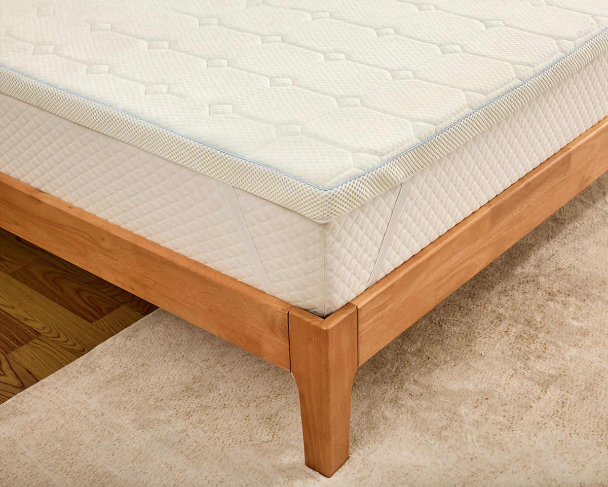 A close-up of a white quilted mattress topper or mattress corner resting on a light wood bed frame over a beige carpet.