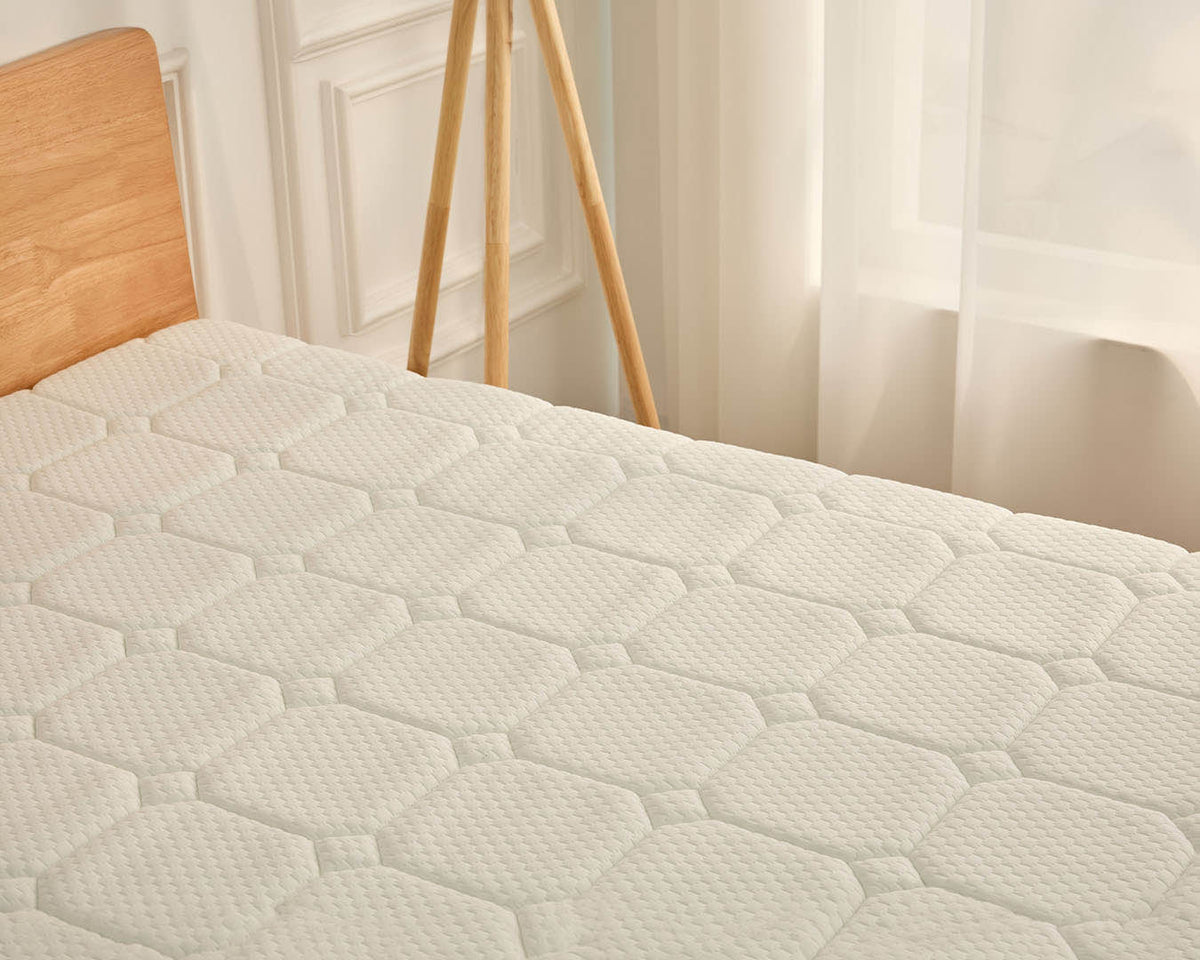 A close-up of a white quilted mattress topper or mattress surface on a bed, with part of a light wood headboard and a wooden tripod stand visible in a softly lit bedroom setting.