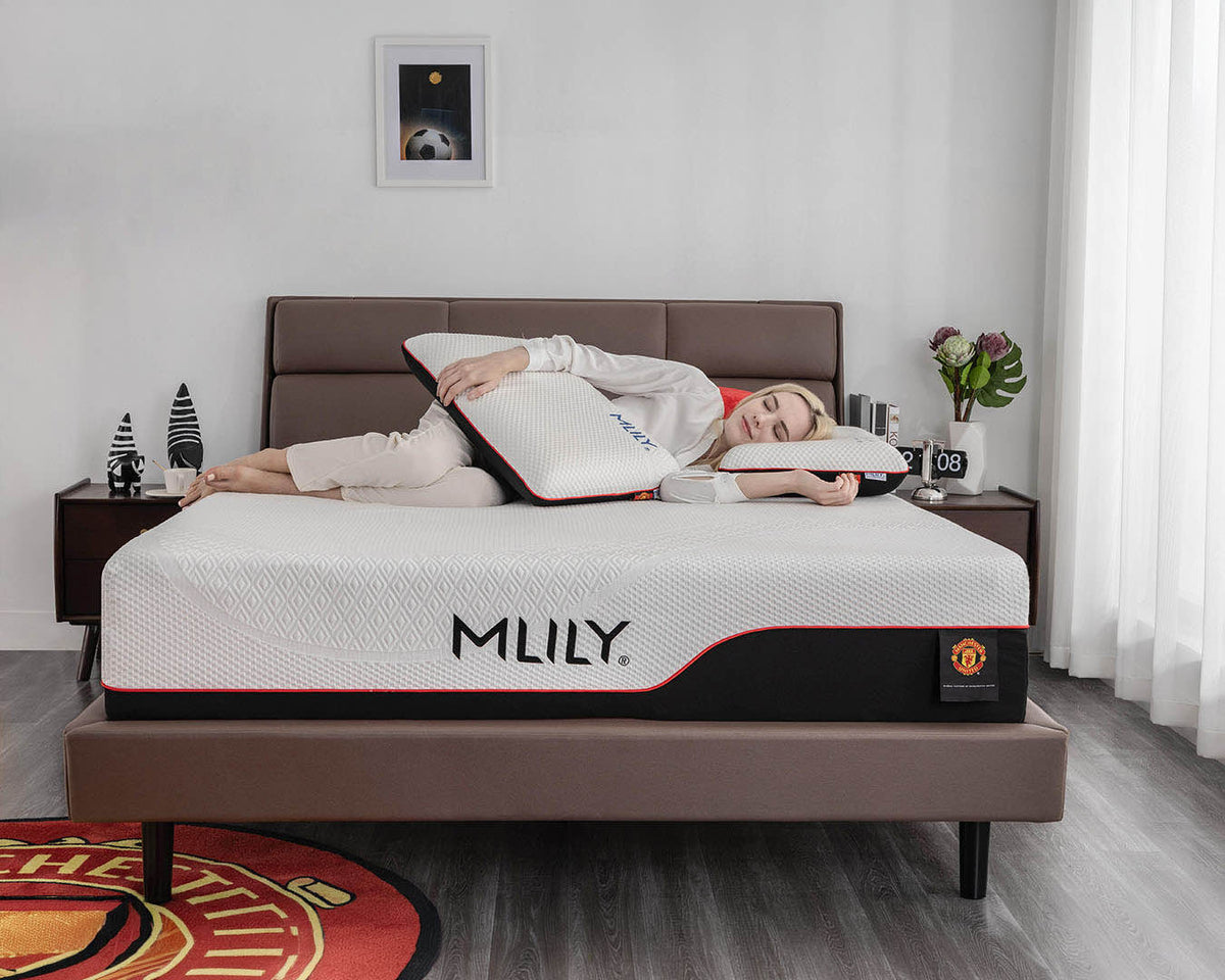 A woman lies on her side on a bed with an MLILY mattress, holding a white pillow. The bedroom has a brown upholstered bed frame, dark nightstands, a window with white curtains, and a framed picture on the wall.