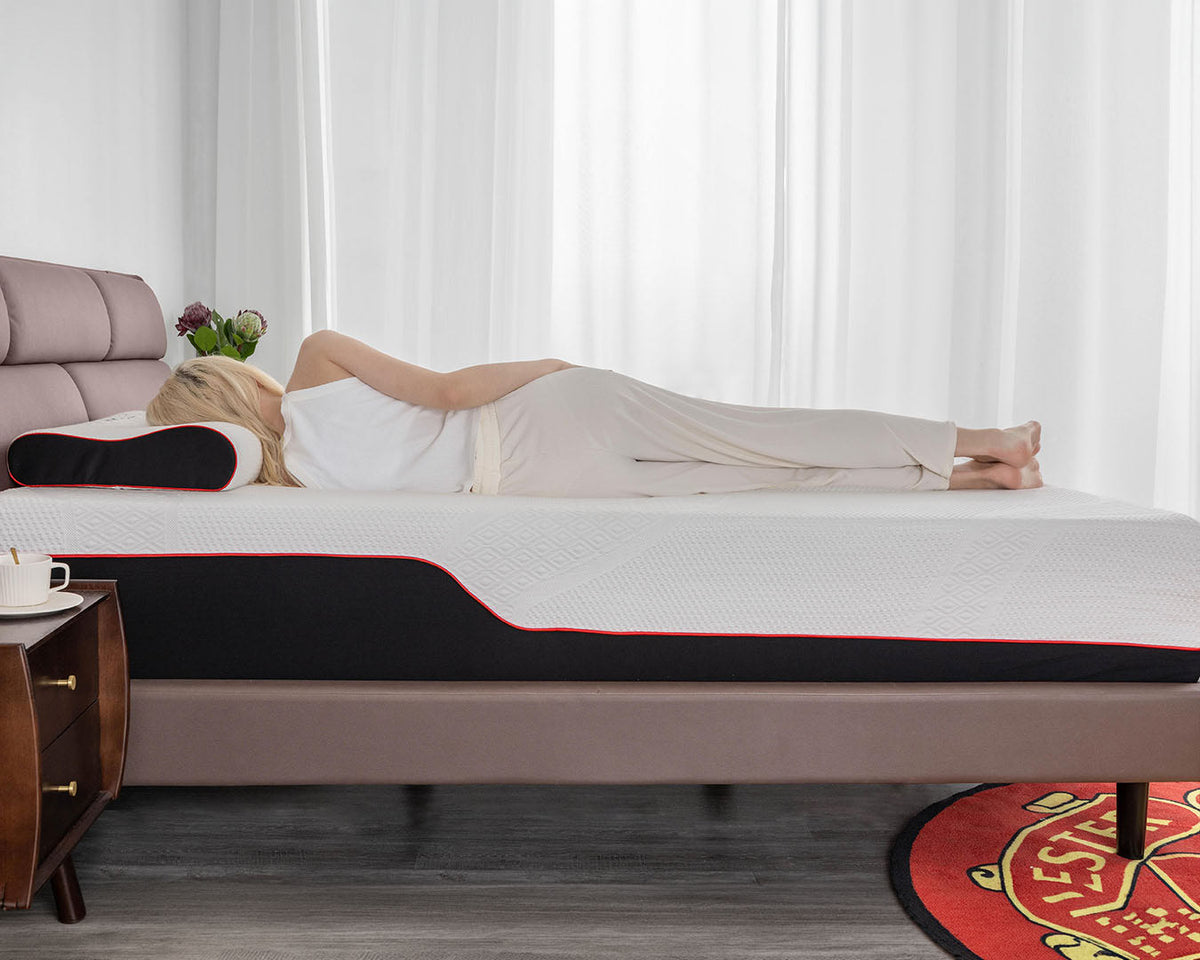 A woman lies on her side on a mattress with a white textured cover, black side panel, and thin red accent line, in a bright bedroom setting.