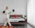 A woman in white pajamas walks beside a bed in a bright bedroom, holding a green book. The bed has a brown upholstered frame, white mattress, and a red plush toy on top.