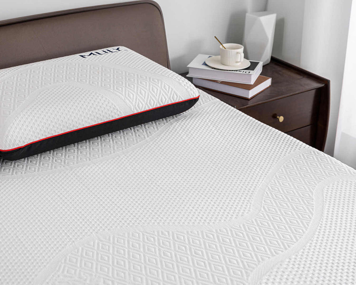 A close-up of a white textured mattress or mattress topper on a bed, with a matching pillow featuring a black and red edge resting against a dark headboard. A wooden nightstand with a cup and books is visible in the background.
